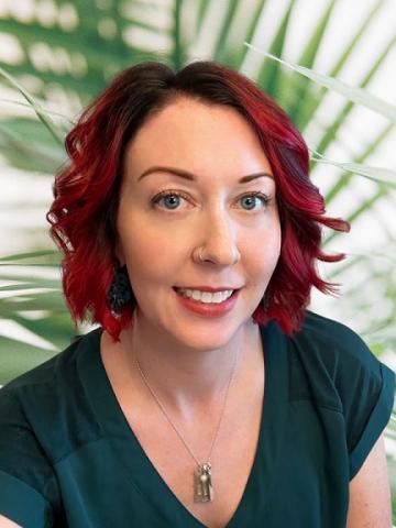 Photo of Dr. Ellen Moore. Palm leaf background, red and black curled hair, smiling, peach skin tone, nose ring, green shirt. Headshot